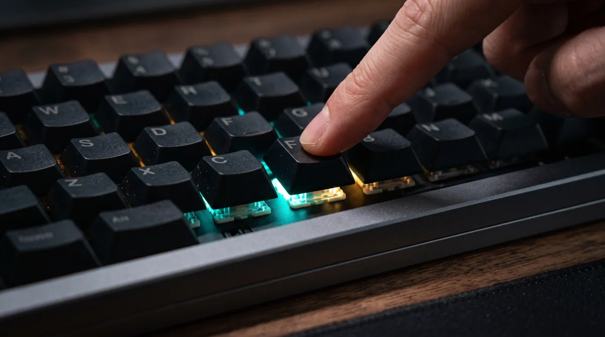 Teclado mecânico preto sobre mesa de madeira, com switches iluminados em verde e âmbar visíveis sob teclas removidas, dedo pressionando uma tecla ao lado